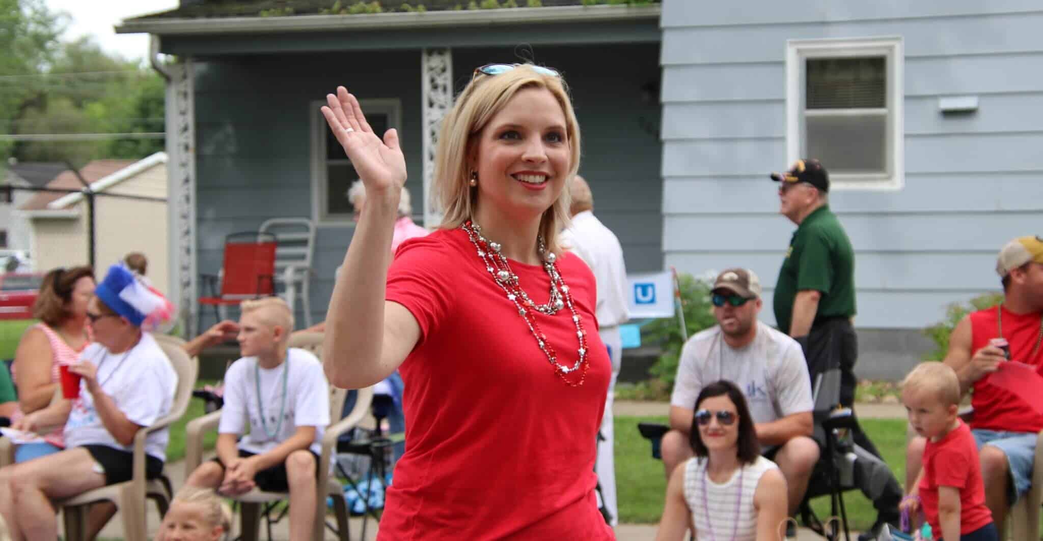 Iowa Congresswoman Ashley Hinson waving and smiling during a parade, with spectators seated in the background.