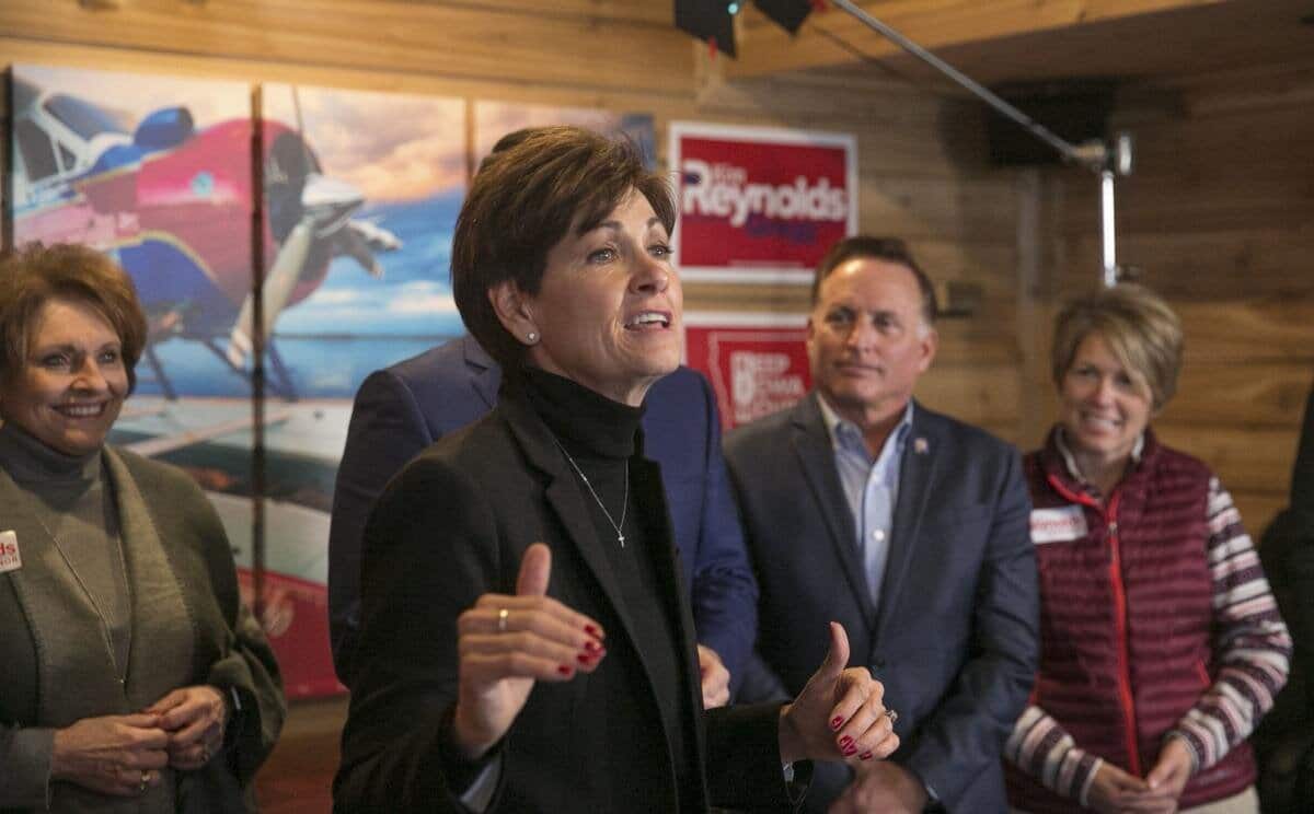 Kim Reynolds speaking at an indoor campaign event, with supporters standing behind her and campaign signs in the background
