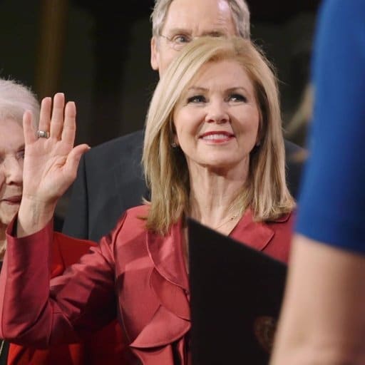 Marsha Blackburn raising her right hand during a swearing-in ceremony, wearing a red suit and smiling.