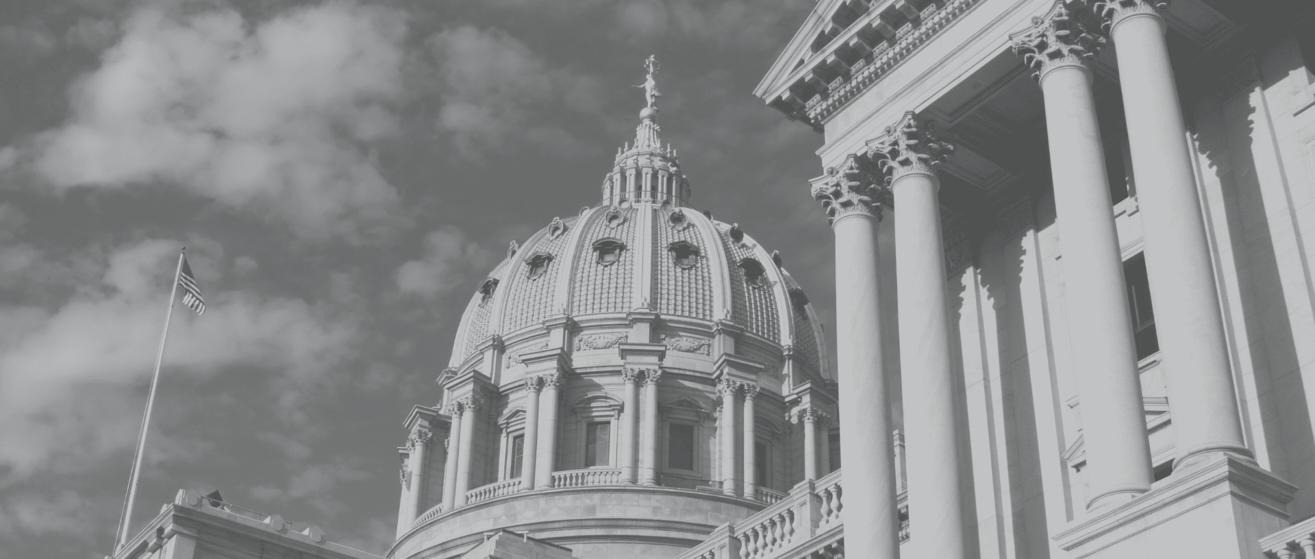 Black and white image of Pennsylvania State Capitol building in Harrisburg, featuring a domed roof and classical columns under a partly cloudy sky.