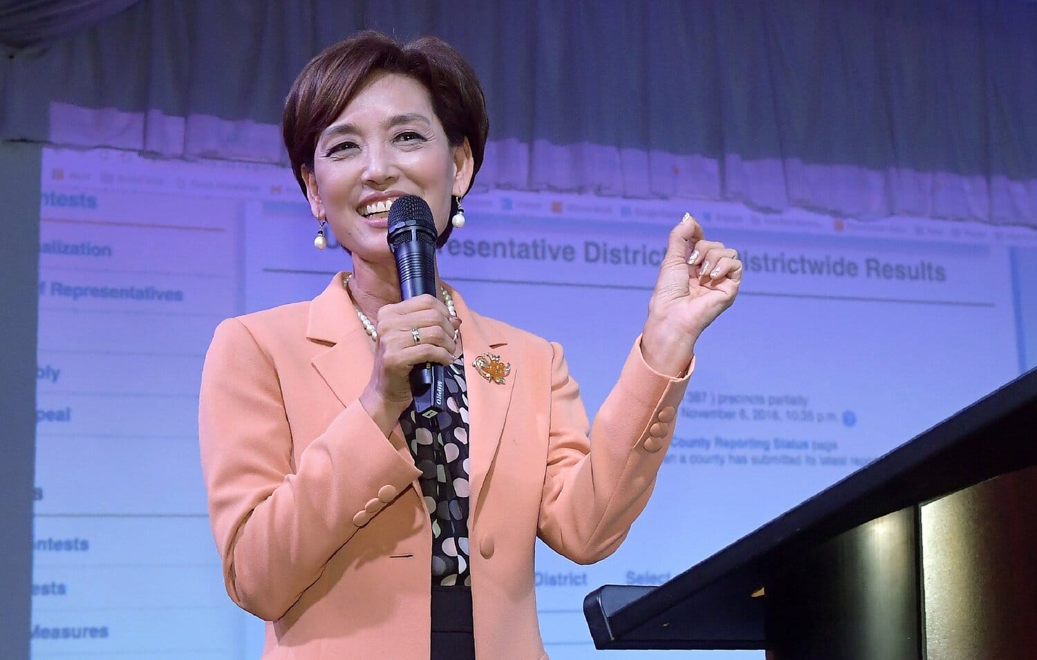 Young Kim speaking into a microphone at a podium while smiling